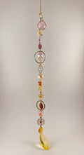 A hanging beaded suncatcher or gingham charm with a gold-tone chain, featuring faceted beads in pink, amber, yellow, and iridescent white, along with decorative metal floral and circular elements, photographed against a plain light beige background.