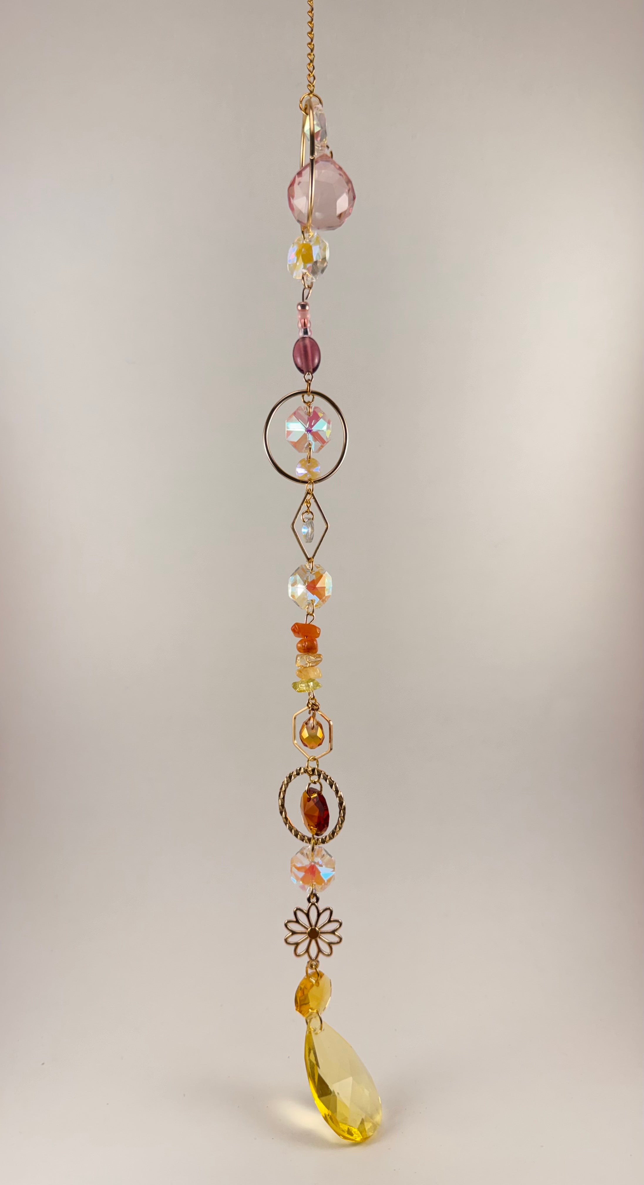 A hanging beaded suncatcher or gingham charm with a gold-tone chain, featuring faceted beads in pink, amber, yellow, and iridescent white, along with decorative metal floral and circular elements, photographed against a plain light beige background.