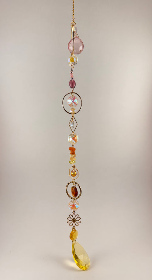 A hanging beaded suncatcher or gingham charm with a gold-tone chain, featuring faceted beads in pink, amber, yellow, and iridescent white, along with decorative metal floral and circular elements, photographed against a plain light beige background.